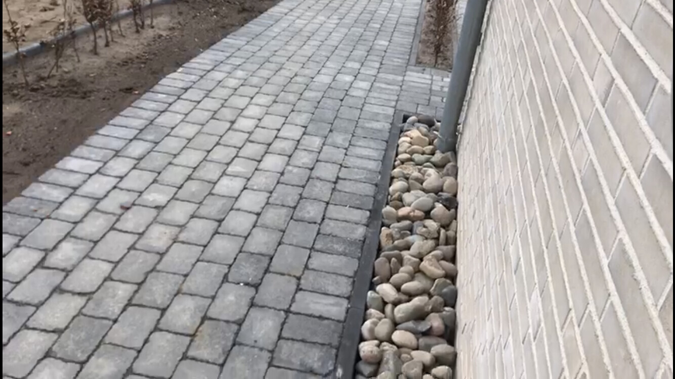 Gepflasterter grauer Gehweg neben Betonwand; Kiesel im seitlichen Randbett.