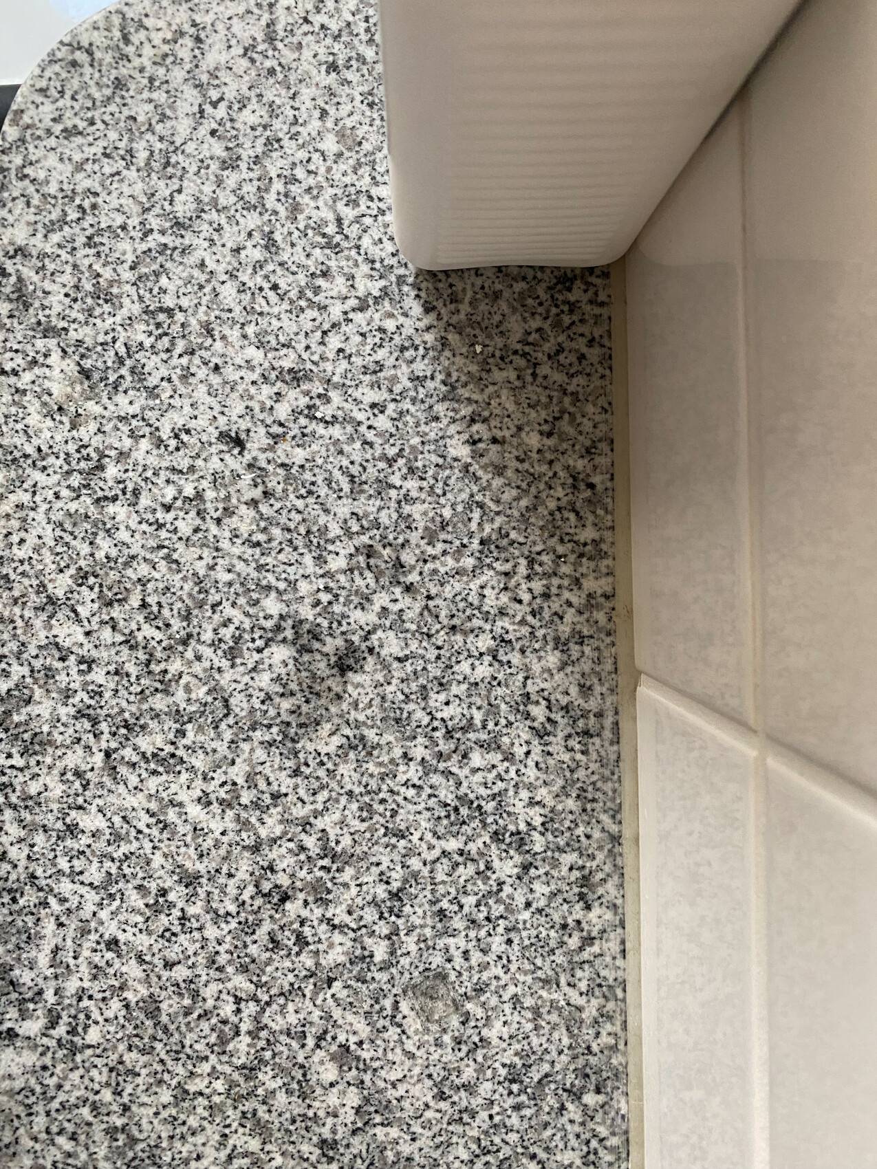 Nahaufnahme einer grauen Granitplatte neben einer beigen Fliesenwand, Ecke eines weißen Möbelstücks.