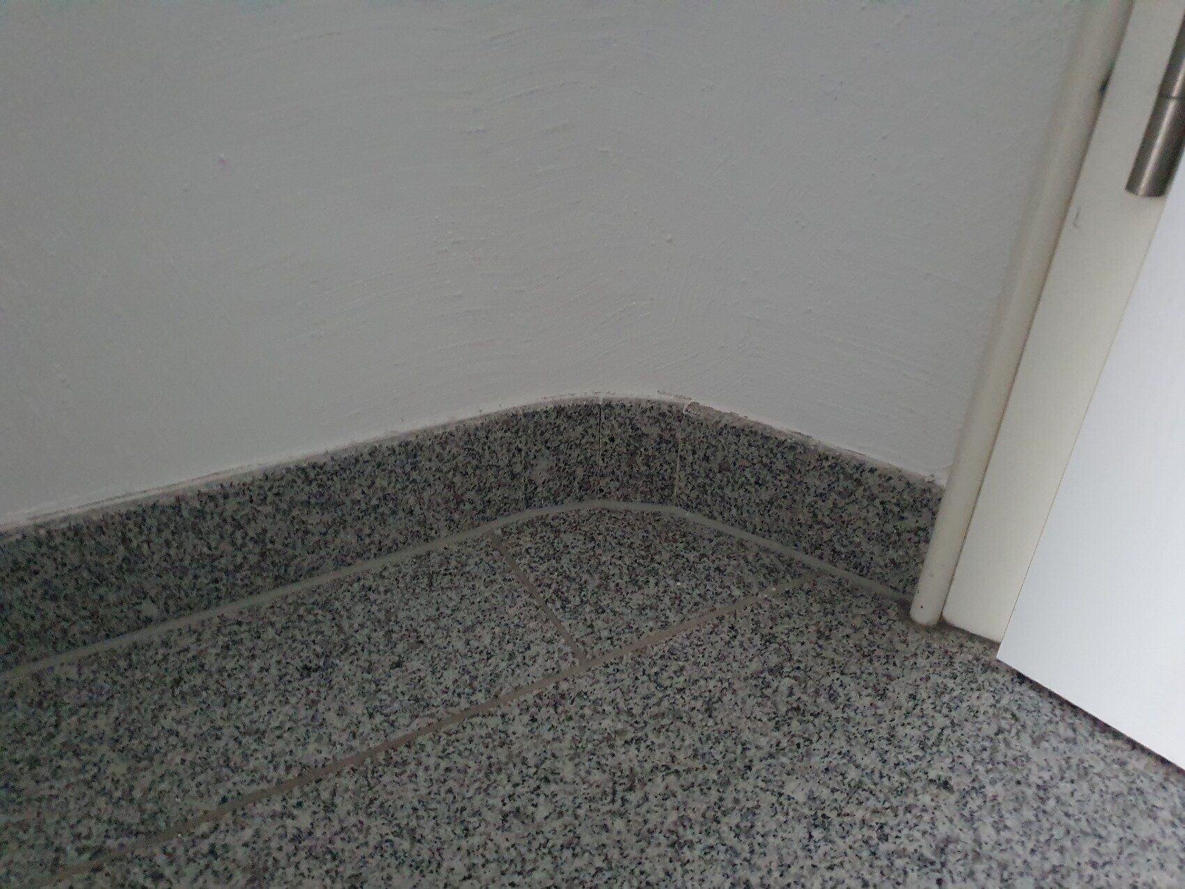 Eckbereich: grauer Granitfliesenboden, weiße Wand, Tür rechts.