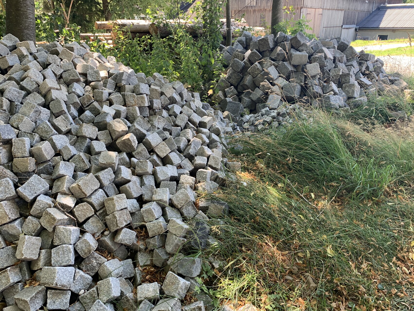 Zwei Haufen grauer Pflastersteine bilden Randmauern neben Gras am Wegesrand, Hintergrund mit Bäumen.