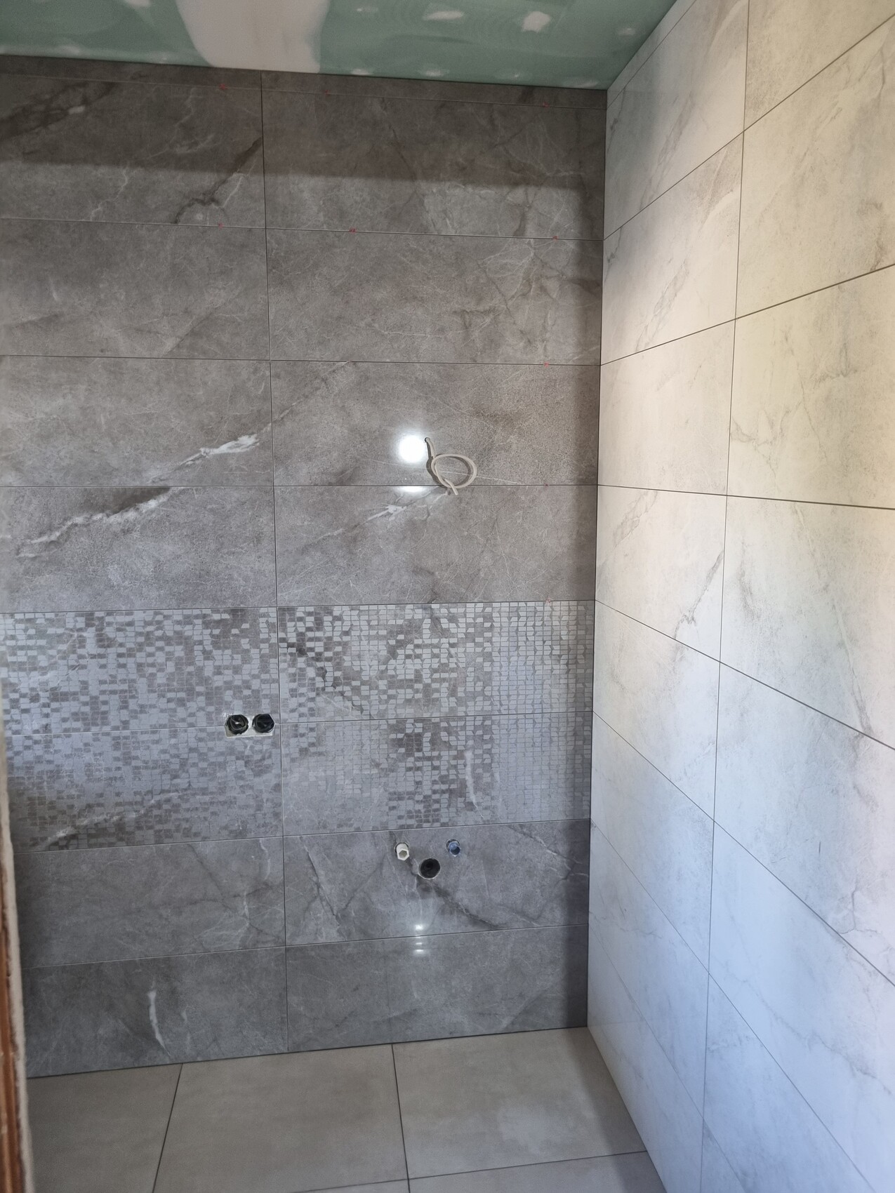 Badezimmerwand aus grauen und weißen Fliesen mit Mosaikband, Wasseranschlüssen und losem Kabel.