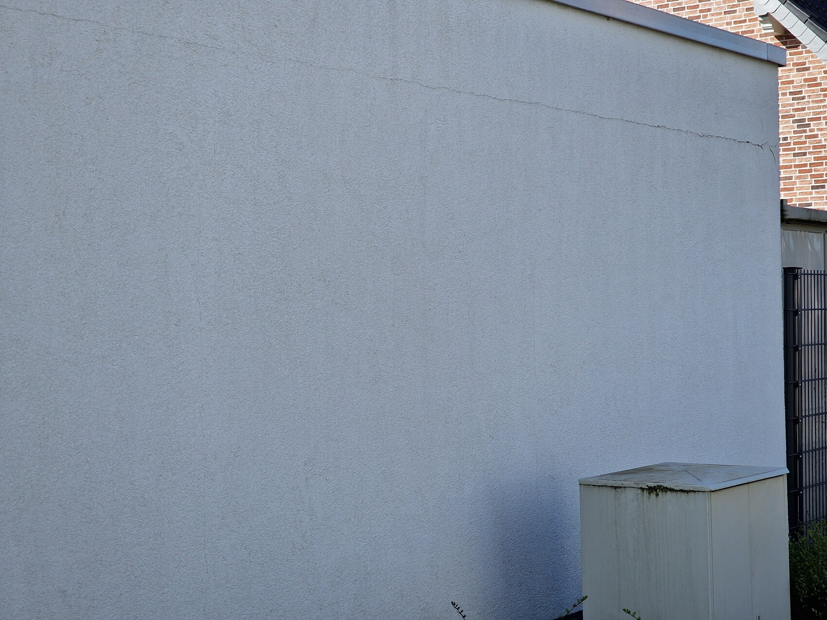 Grau verputzte Außenwand mit sichtbarer Textur; rechts ein Metallzaun und ein kleiner Schrank.