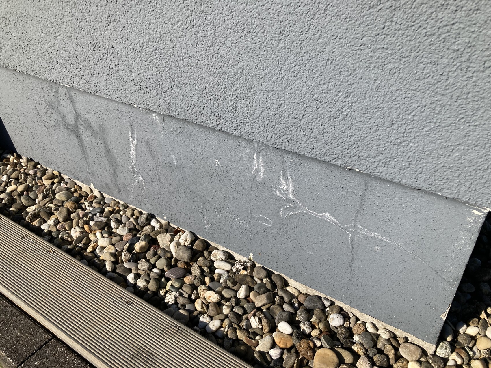 Schräg an einer rauen grauen Wand lehnt eine graue Platte; davor Kieselsteine.