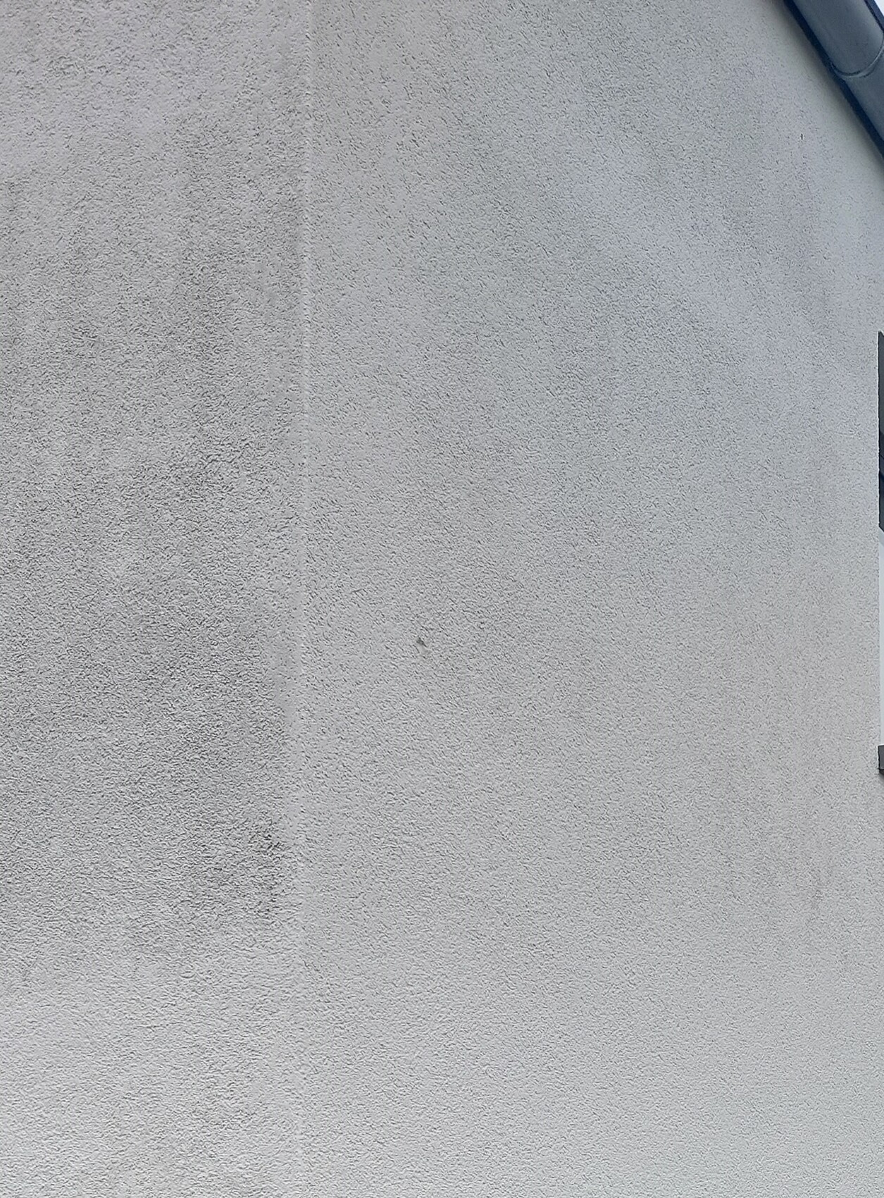 Helle, rauhe Putzwand mit Struktur; oben rechts ein Rohr bzw. Dachrinnen-Detail erkennbar.