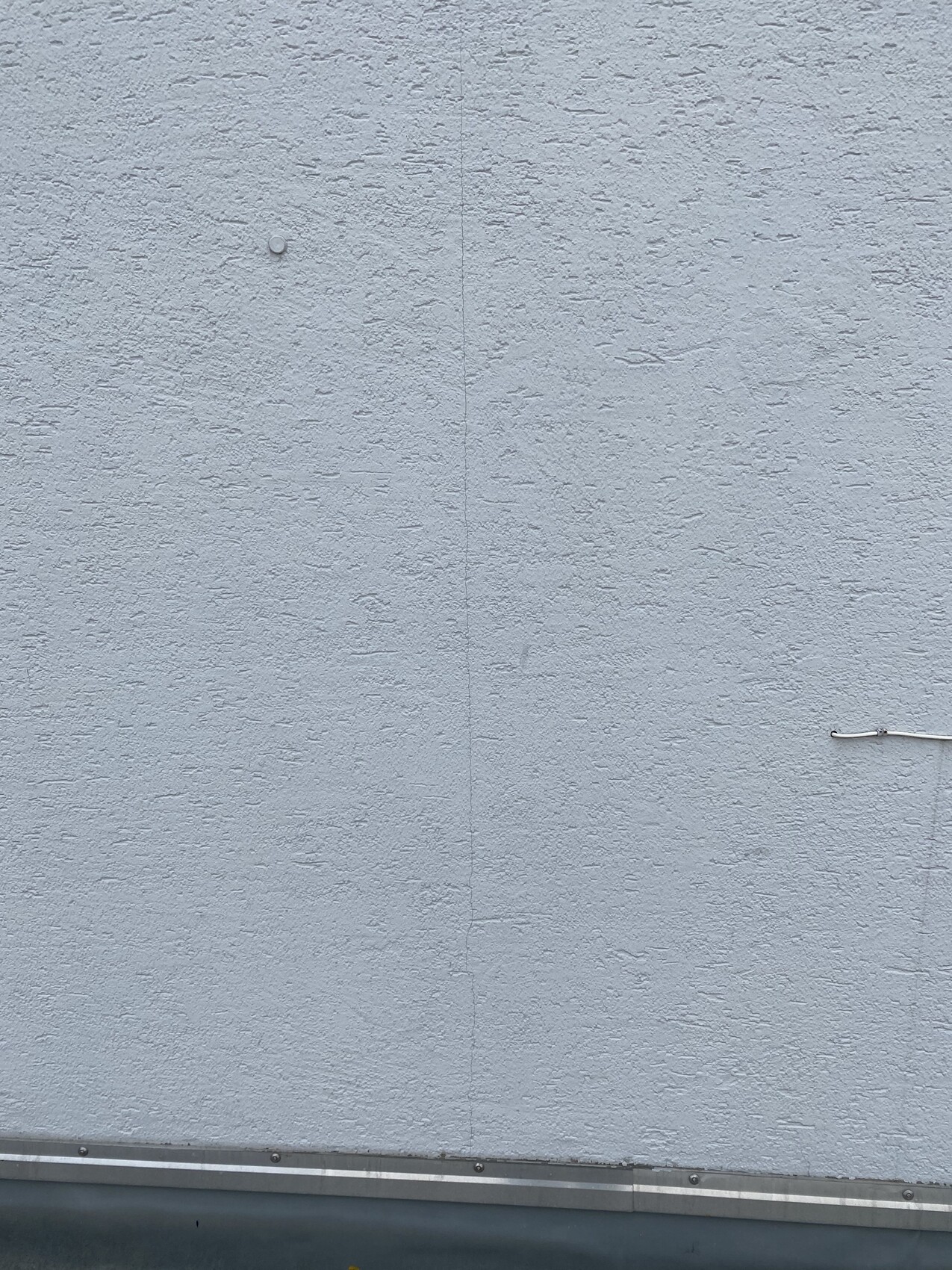 Helle verputzte Wand mit Textur, vertikale Fuge, kleinem Knopf, Kabel am Rand, Metallleiste unten