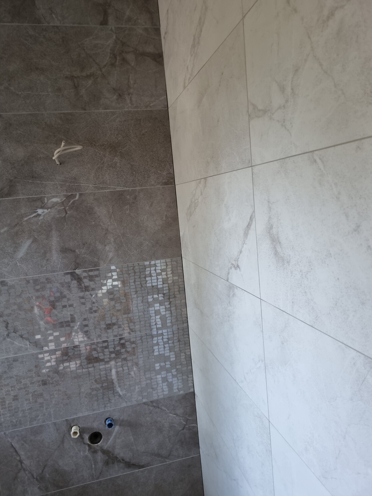 Unfertige Badezimmerwand mit dunklen Fliesen, Mosaikband und hellen Fliesen; Rohre sichtbar.