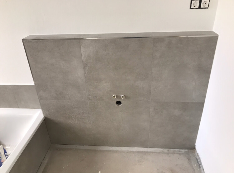 Graue Fliesenwand im Badezimmer mit zwei Wasseranschlüssen und Abfluss.