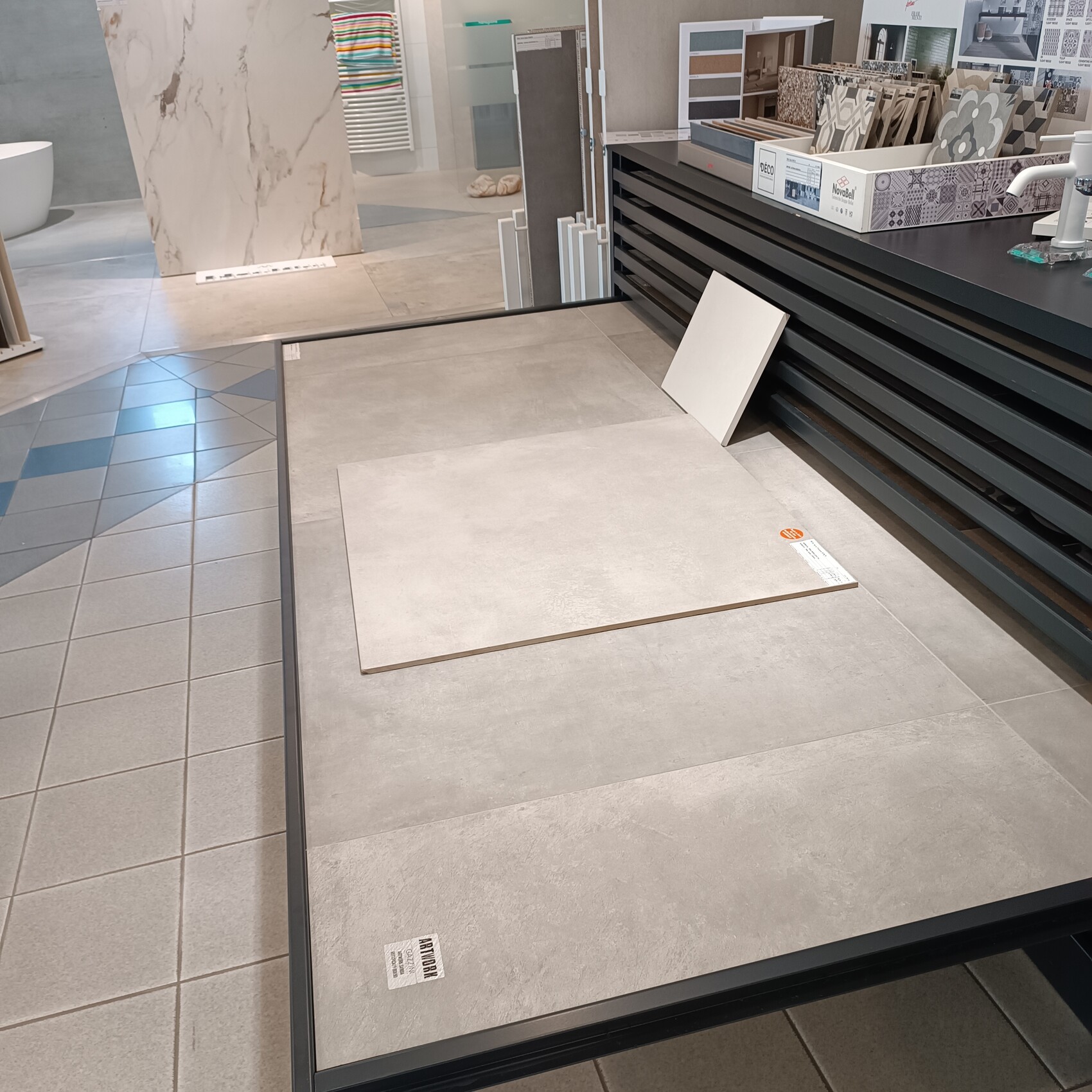Fliesenstudio: Großer Tisch mit hellgrauen Bodenfliesenplatten, rechts Theke mit Musterkacheln.