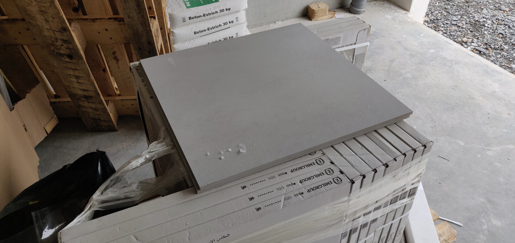 Eine große graue Bauplatte liegt schräg auf einem Palettenstapel; dahinter Beton-Estrich-Säcke.