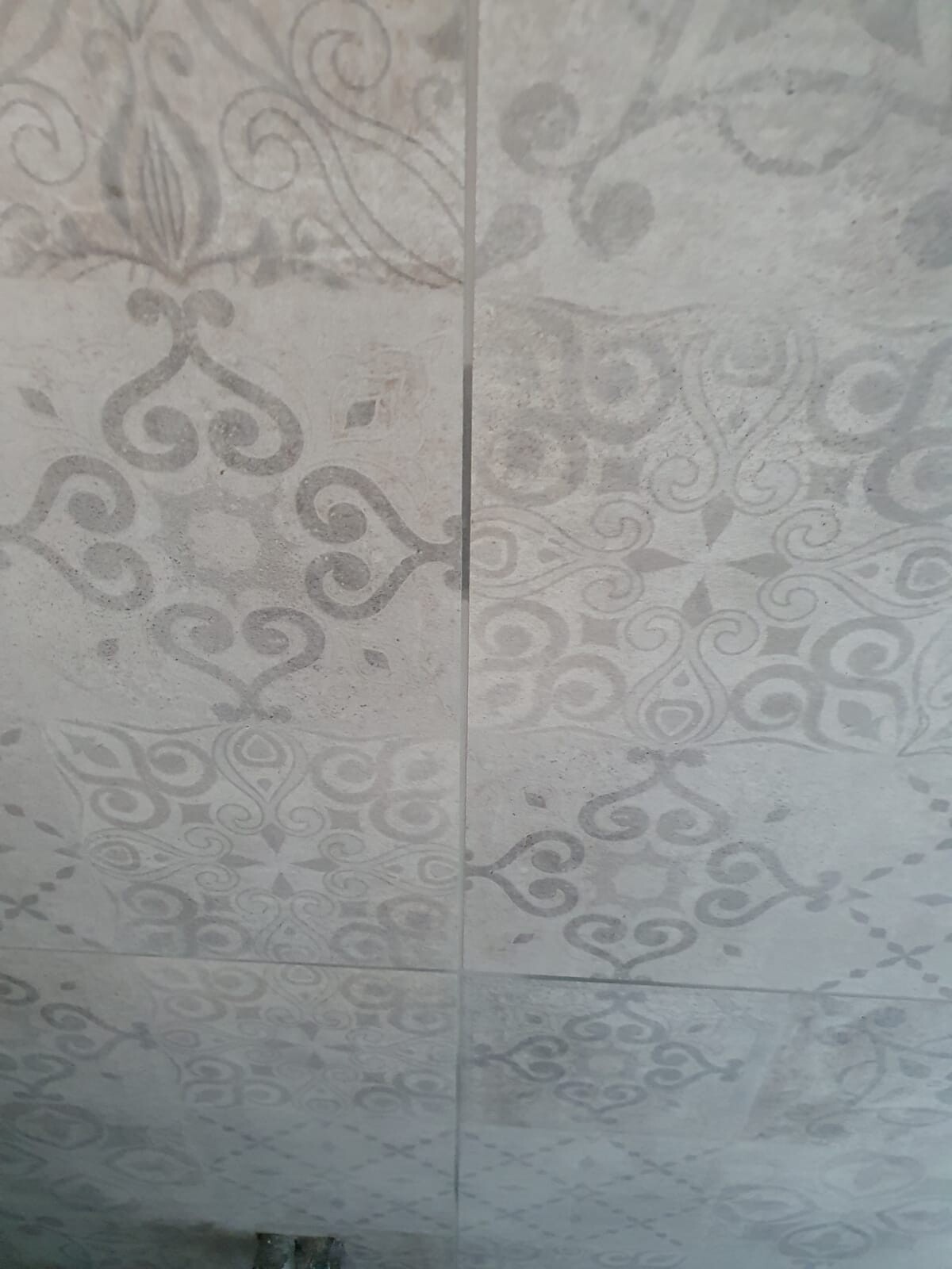 Hellgraue Fliesenwand mit symmetrischen, ornamentalen Rosettenmustern in Graustufen.