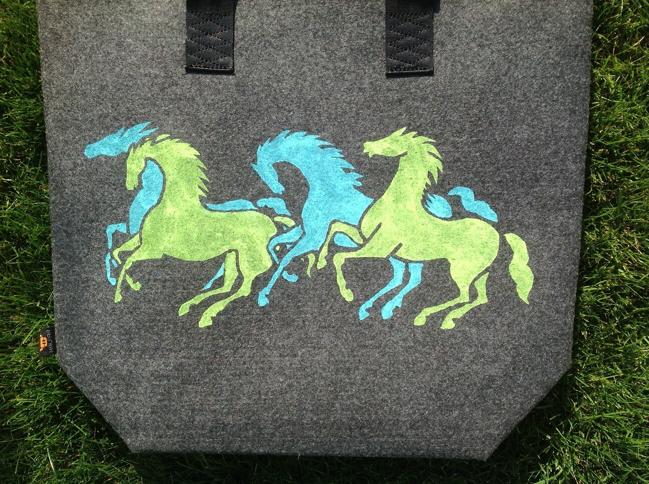 Grauer Stoffbeutel mit drei galoppierenden Pferden in Blau und Grün auf Gras.