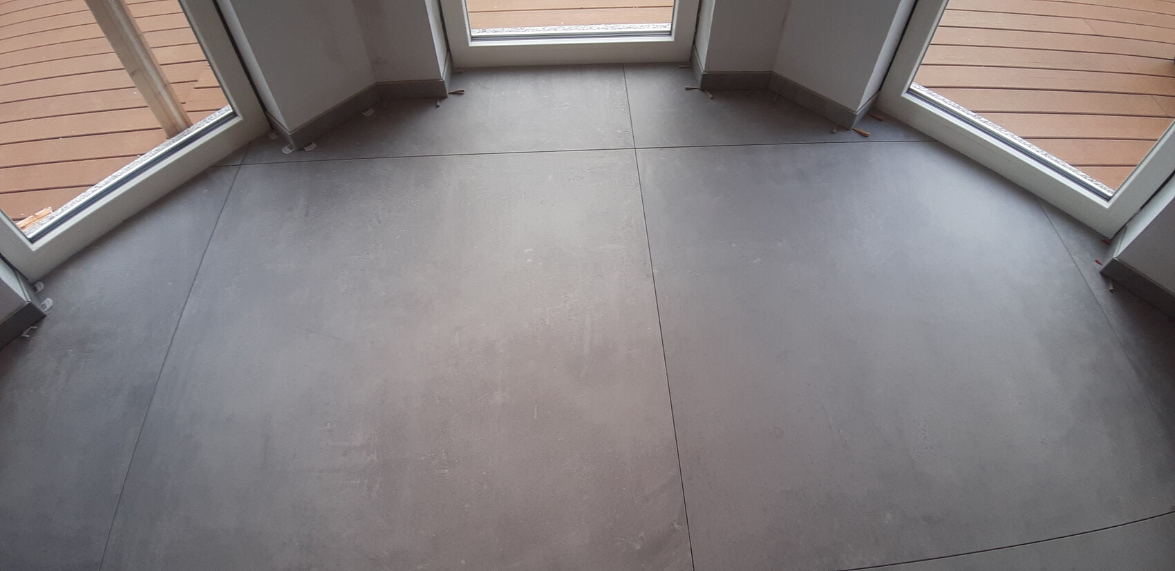 Innenraum mit grauen Bodenplatten und bodentiefen Glasfenstern, Blick auf eine Holzterrasse.