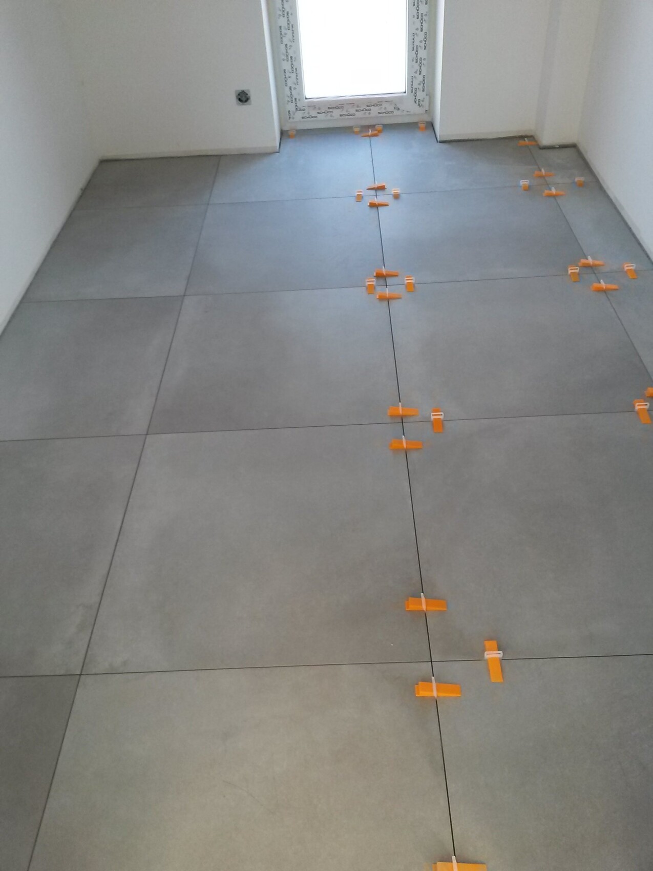 Verlegter Boden: graue Fliesen, orange Fliesenkeile in den Fugen, Tür am Ende des Raums.