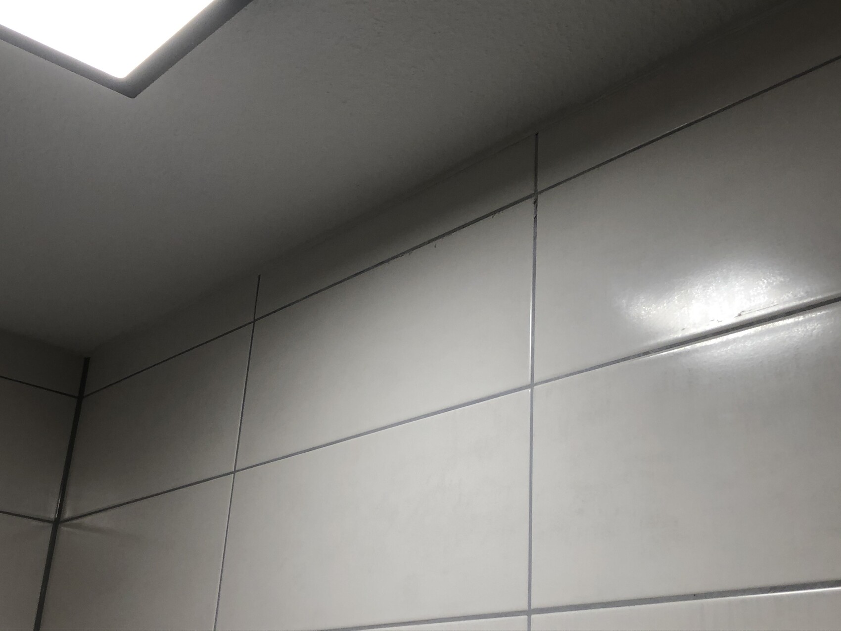 Eckige, glänzende graue Fliesenwand mit dunklen Fugen; helle Deckenleuchte oben links.