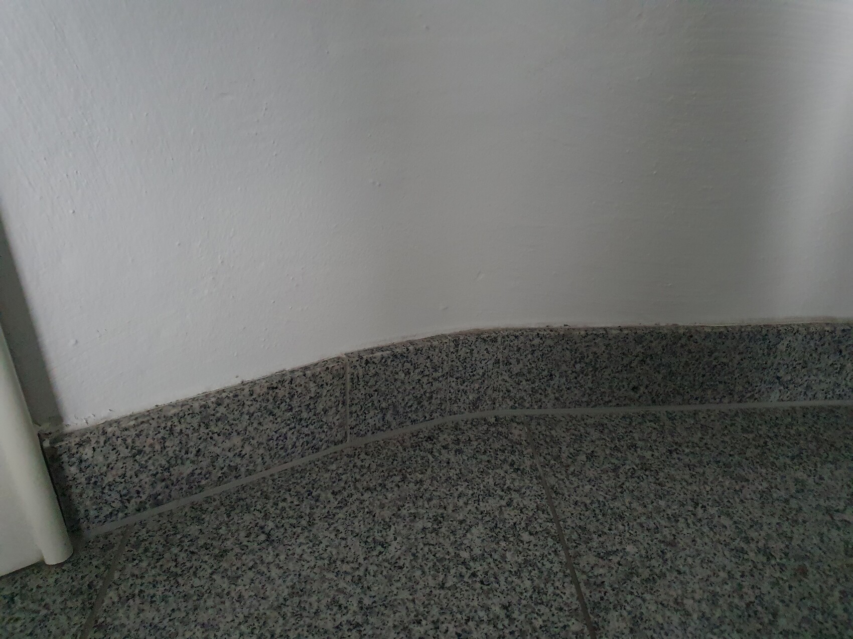 Weiße Wand trifft auf graue gesprenkelte Bodenfliesen; runde Kante am unteren Rand der Wand.