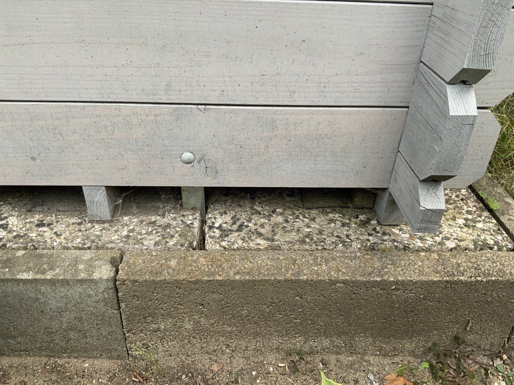 Graue Holzbretter ruhen auf Betonblöcken über einer Betonplatte; Boden mit Kies.