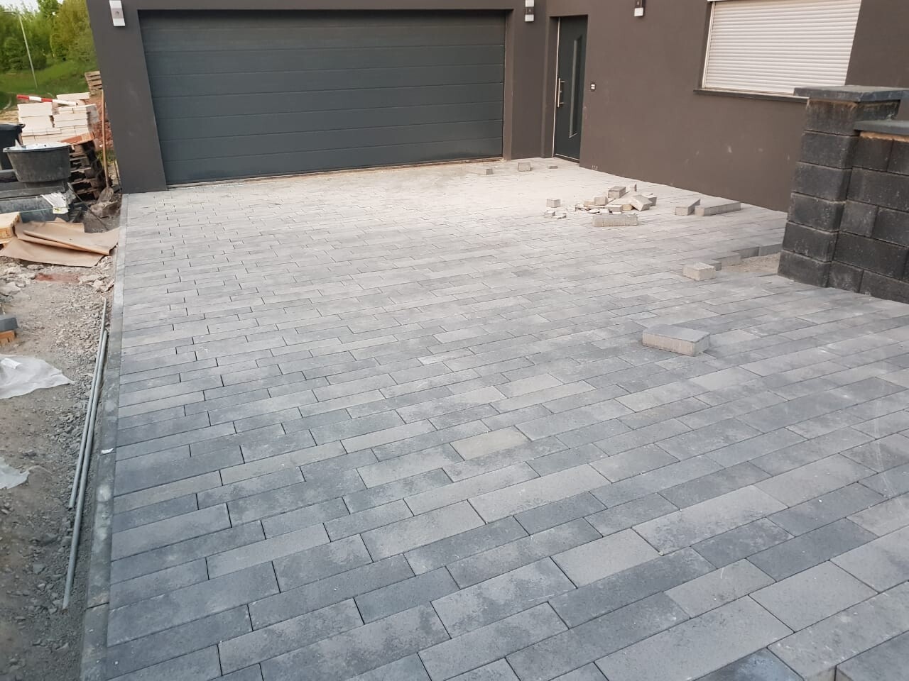 Neu gepflasterte graue Einfahrt vor der Garage und dem Haus; Bau-Materialien links.