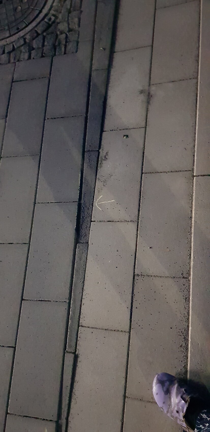 Gehweg aus grauen Pflastersteinen, Pfeilmarkierung, Mannloch links oben, lila Schuh rechts unten.