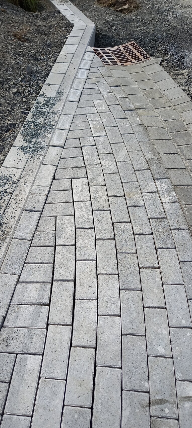 Gepflasterter Gehweg aus rechteckigen Betonpflastersteinen mit Metallrinnenabdeckung am Rand.
