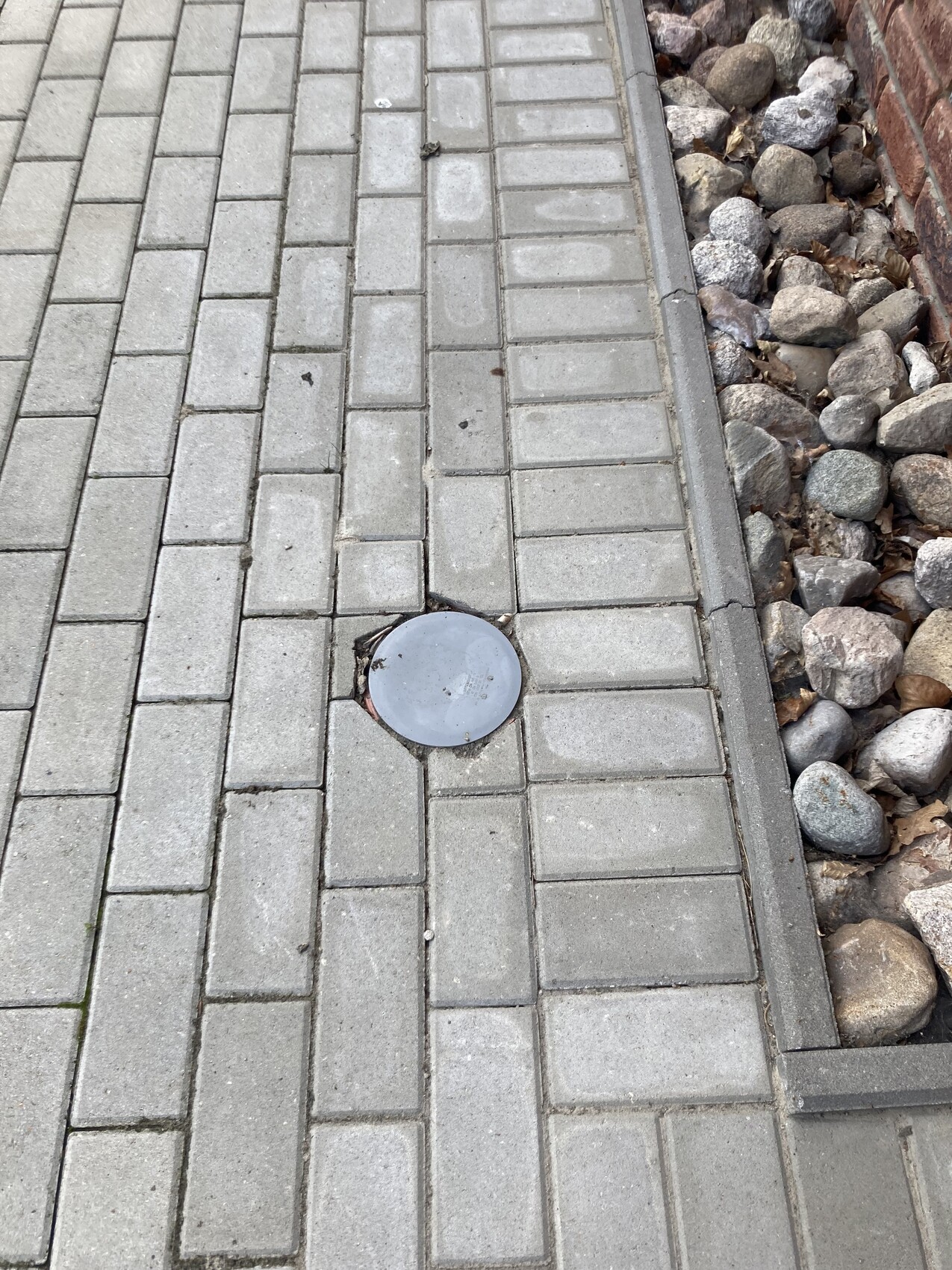 Gehweg aus grauen rechteckigen Pflastersteinen mit rundem Metalldeckel; rechte Seite mit Kieselsteinen.