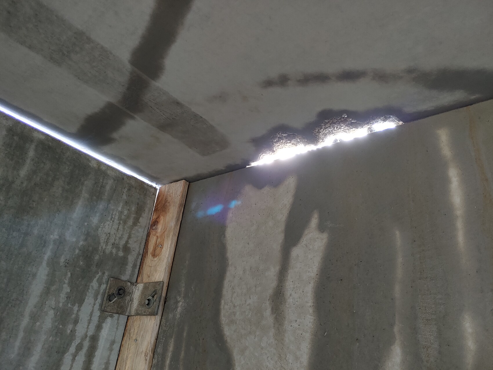 Lichtspalt zwischen Decke und Betonwand; Holzlatte mit Metallbeschlag, Schatten der Kamera sichtbar