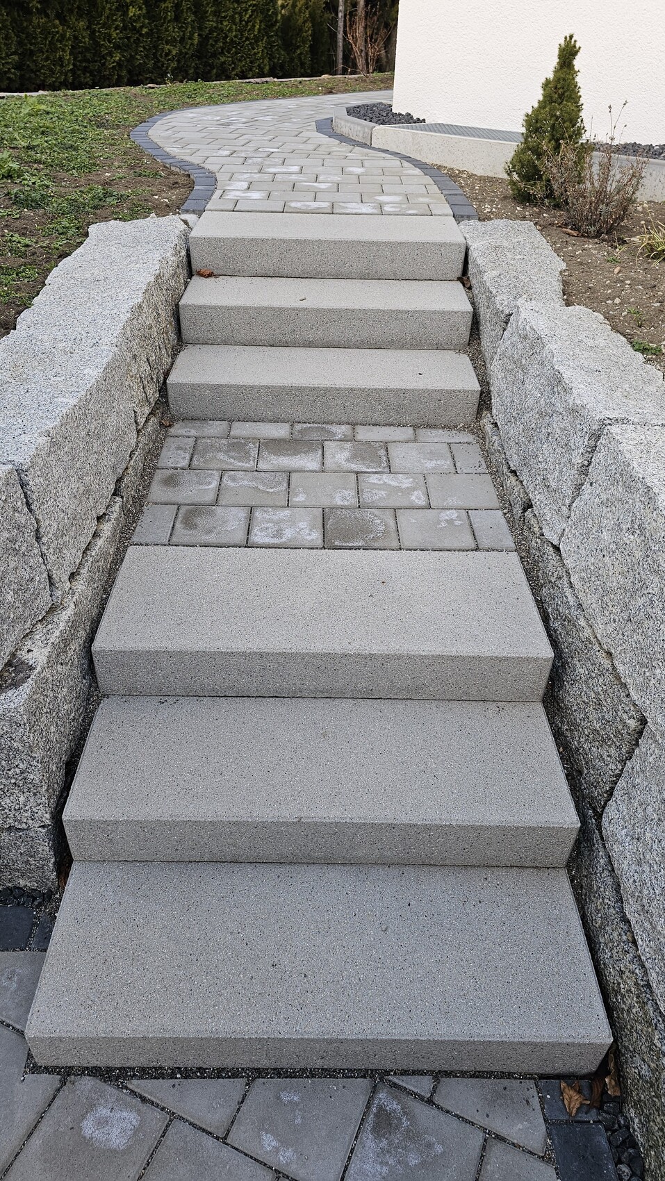 Grauer Betonstufenpfad neben Granitmauer, führt zu einem geschwungenen gepflasterten Weg.