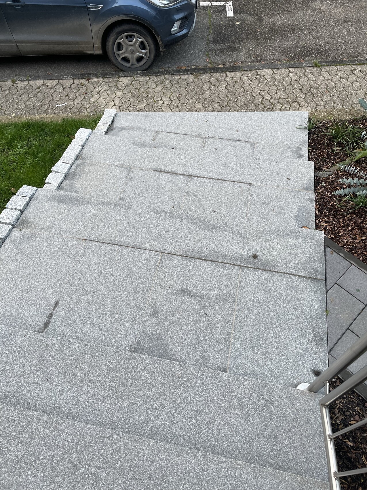 Graue Stein-Treppen mit Geländer führen nach unten; Straße und geparktes Auto im Hintergrund.
