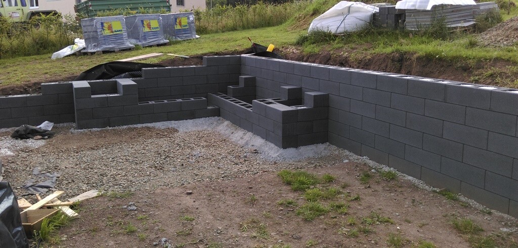 Graue Betonblöcke bilden Fundament- und Innenwaende in einer Baugrube auf Kiesboden.