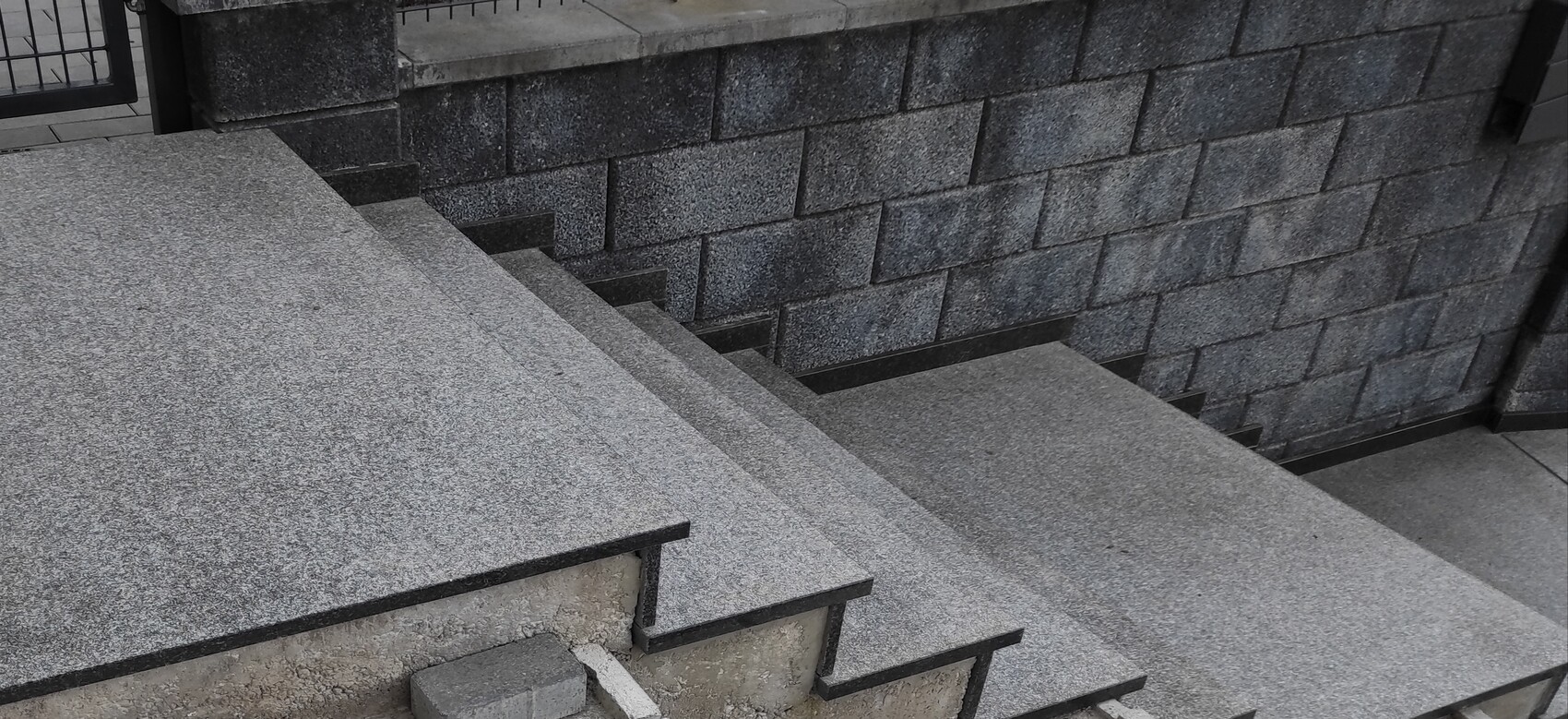 Außenstufen aus grauem Granit führen hinab, rechts eine blockige Betonsteinmauer.