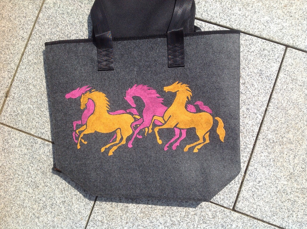 Dunkelgraue Stofftasche mit drei galoppierenden Pferden in Gelb und Pink; schwarze Henkel