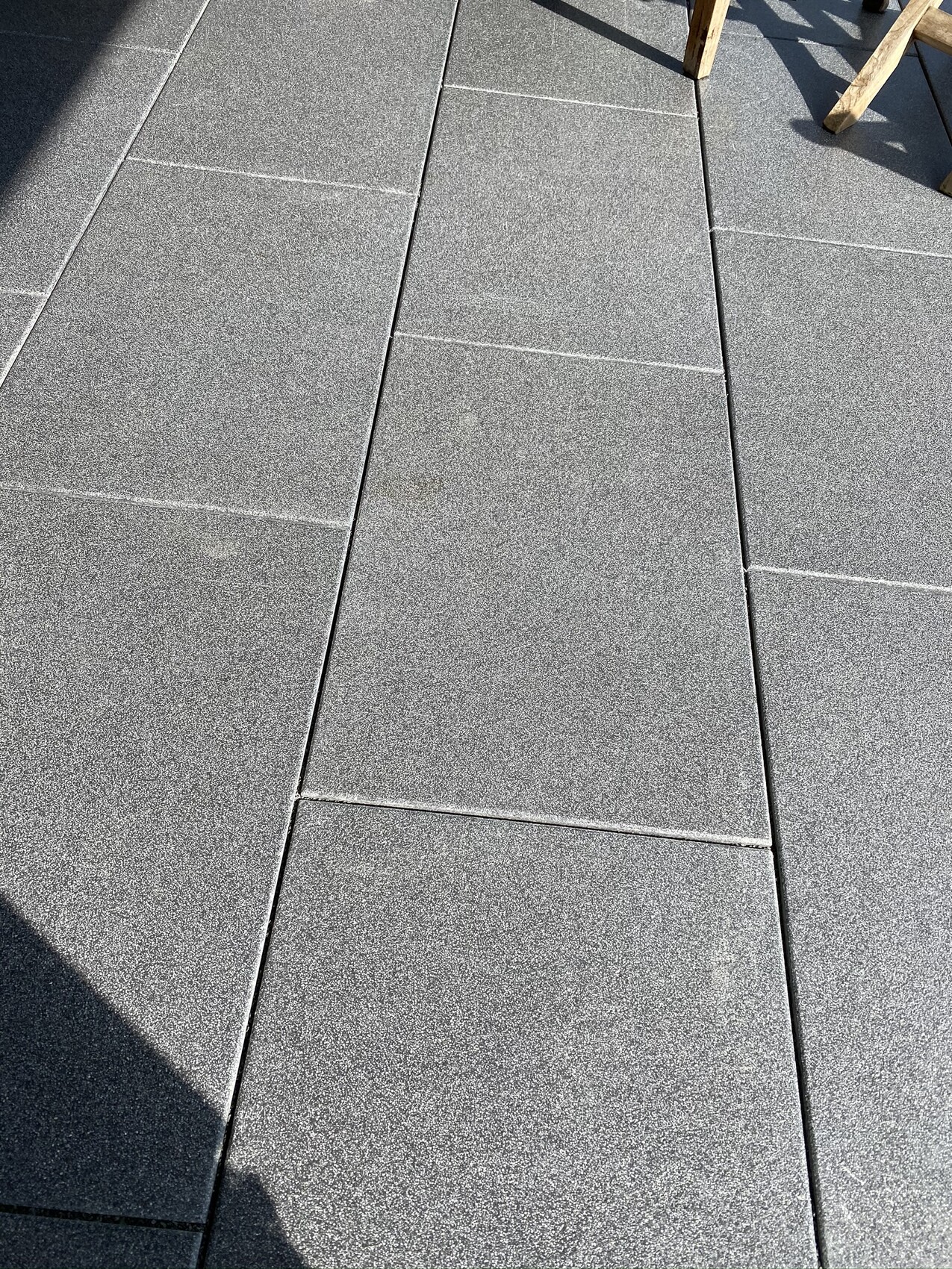Grau gepflasterte, rechteckige Betonplatten als Bodenbelag; Schatten und Holzstuhl-Beine oben rechts.