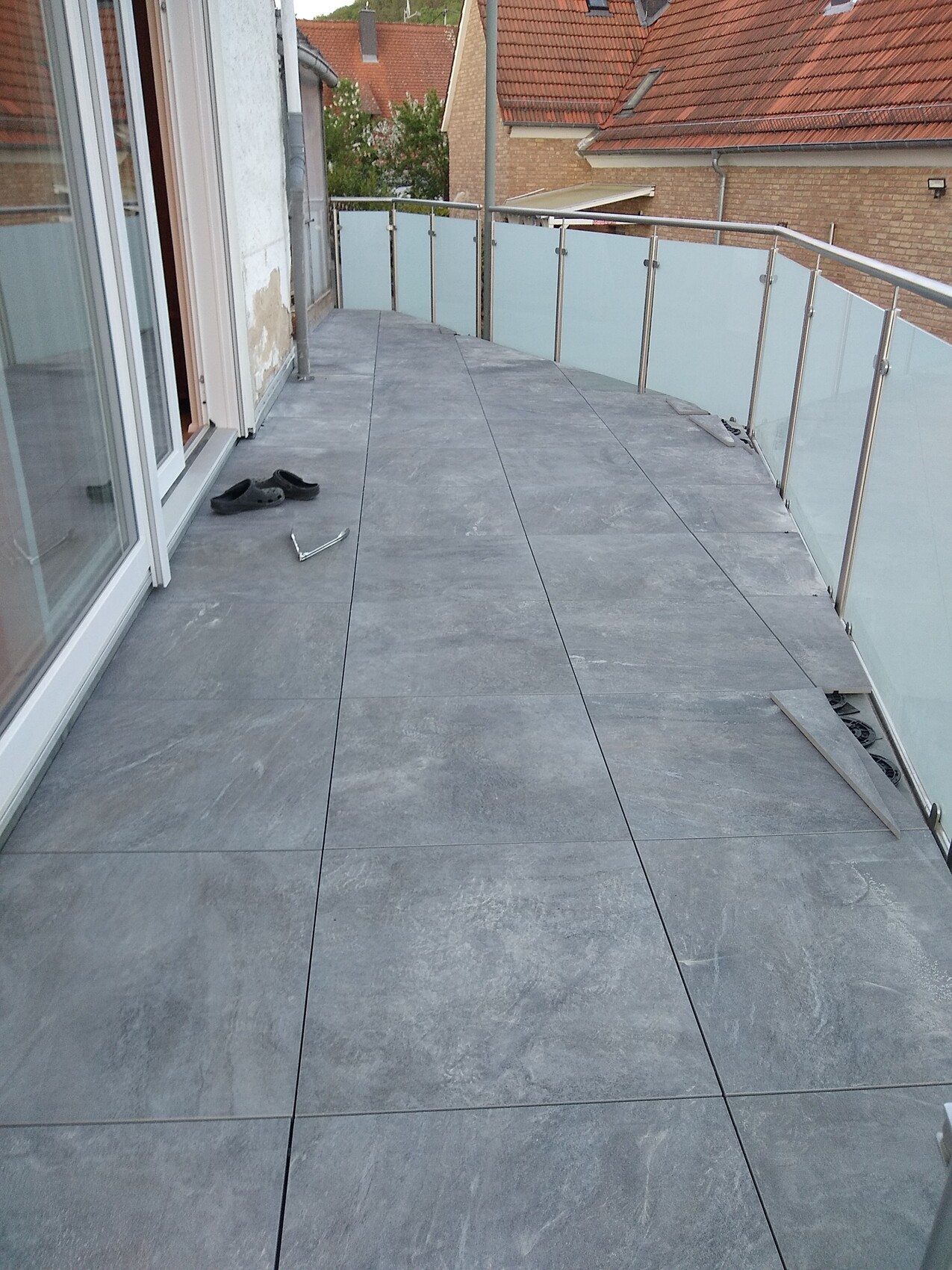 Balkon mit grauen Fliesen, Glasgeländer; Schuhe und Werkzeug liegen auf dem Boden.