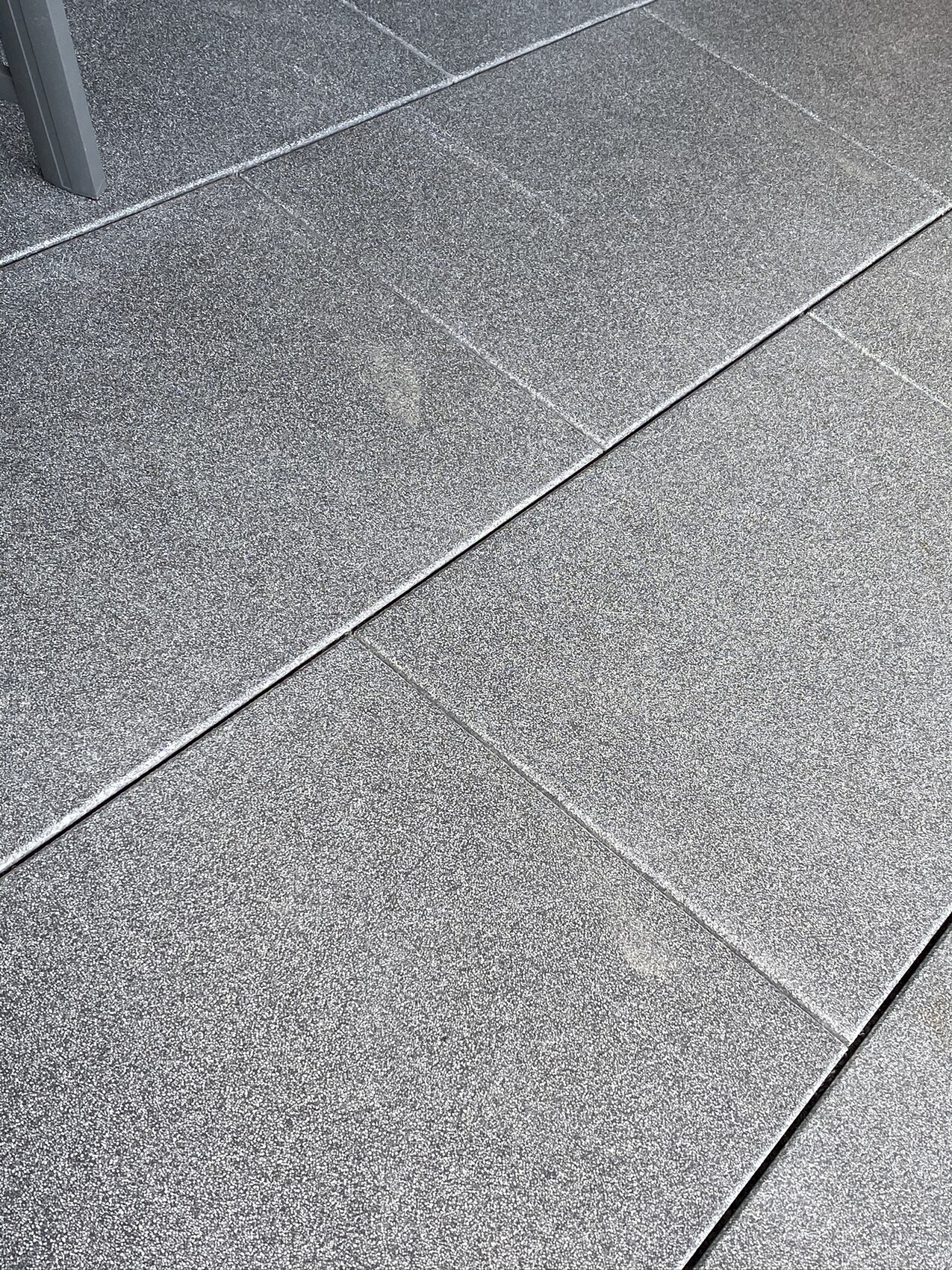 Grau texturierte Terrassenplatten mit Fugen, ein Möbelbein im oberen linken Bereich.