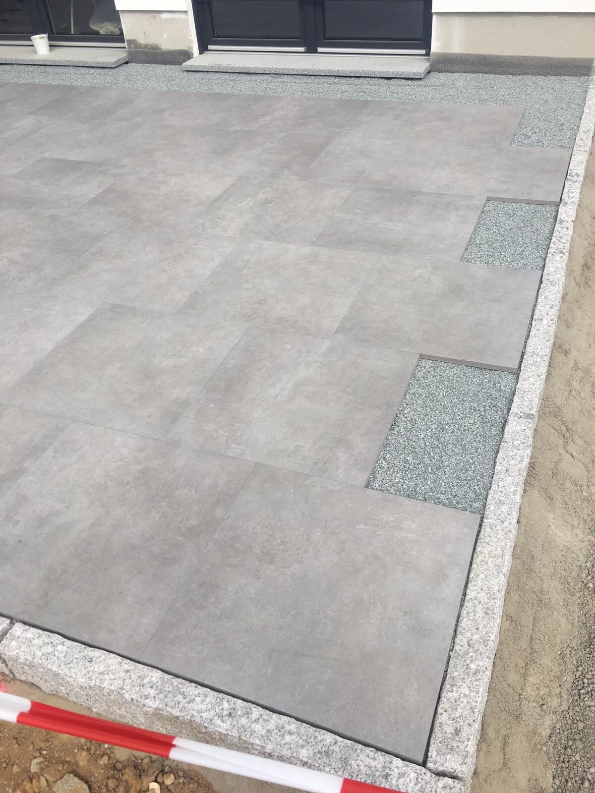 Große graue Terrassenplatten aus Beton, Rand aus Kies neben Haus mit Tür und Fensterfront.