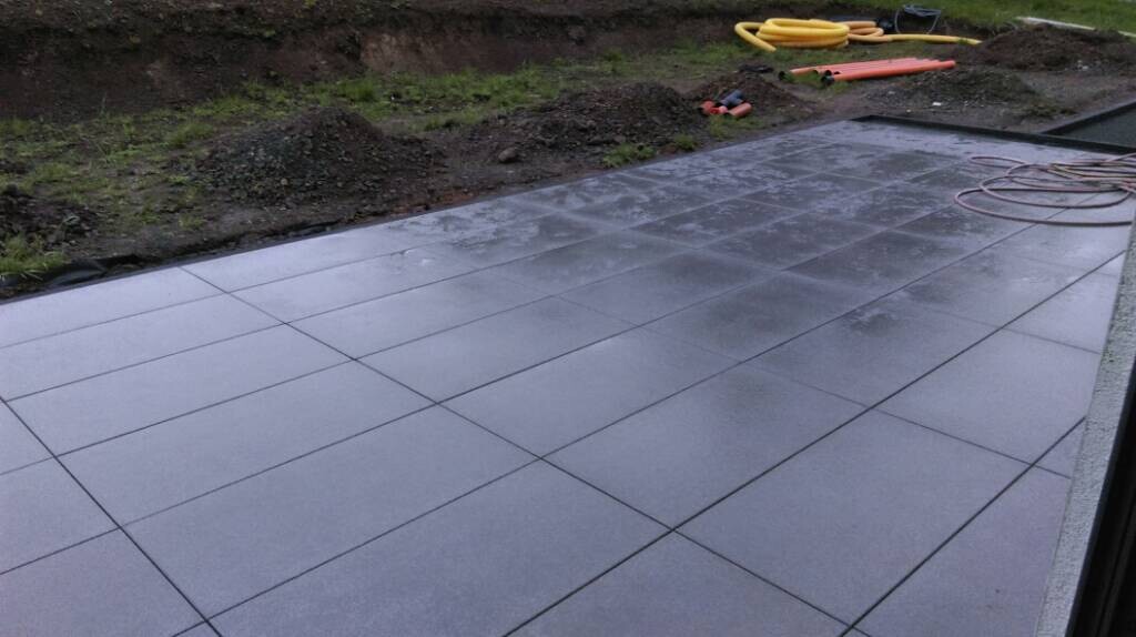 Nass glänzende graue Terrassenplatten auf Terrasse; Bauwerkzeuge und Schlauch im Hintergrund.