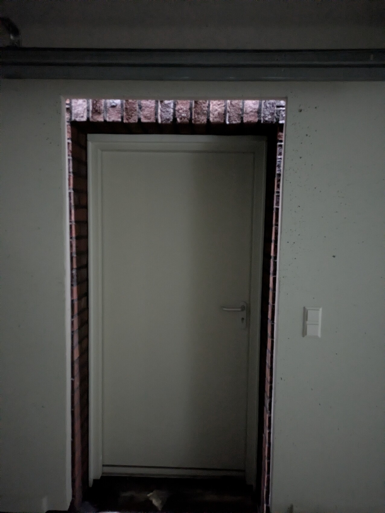 Graue Tür mit Backsteinrahmen im dunklen Flur, Innenbereich sichtbar