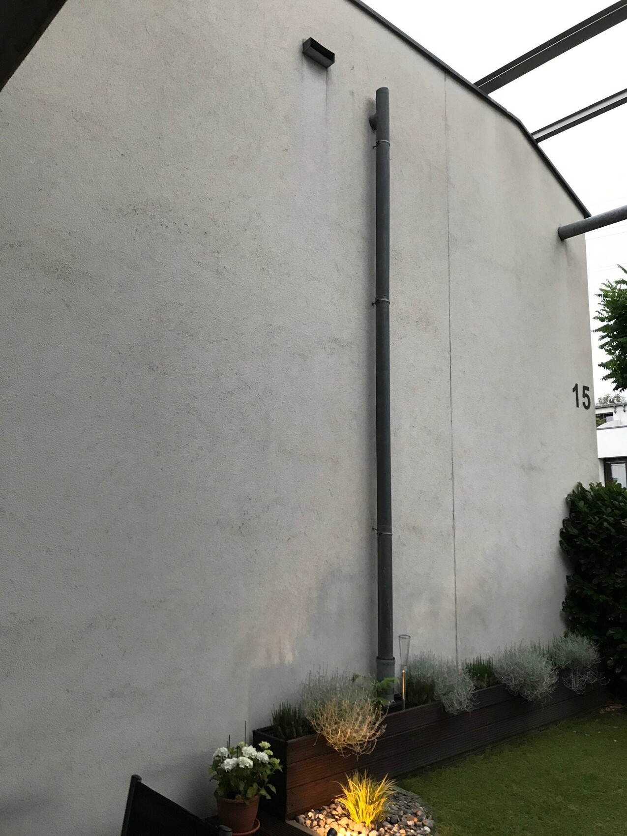 Graue Außenwand mit senkrechtem Regenrohr; Gartenbeet mit Pflanzen unten, Hausnummer 15.