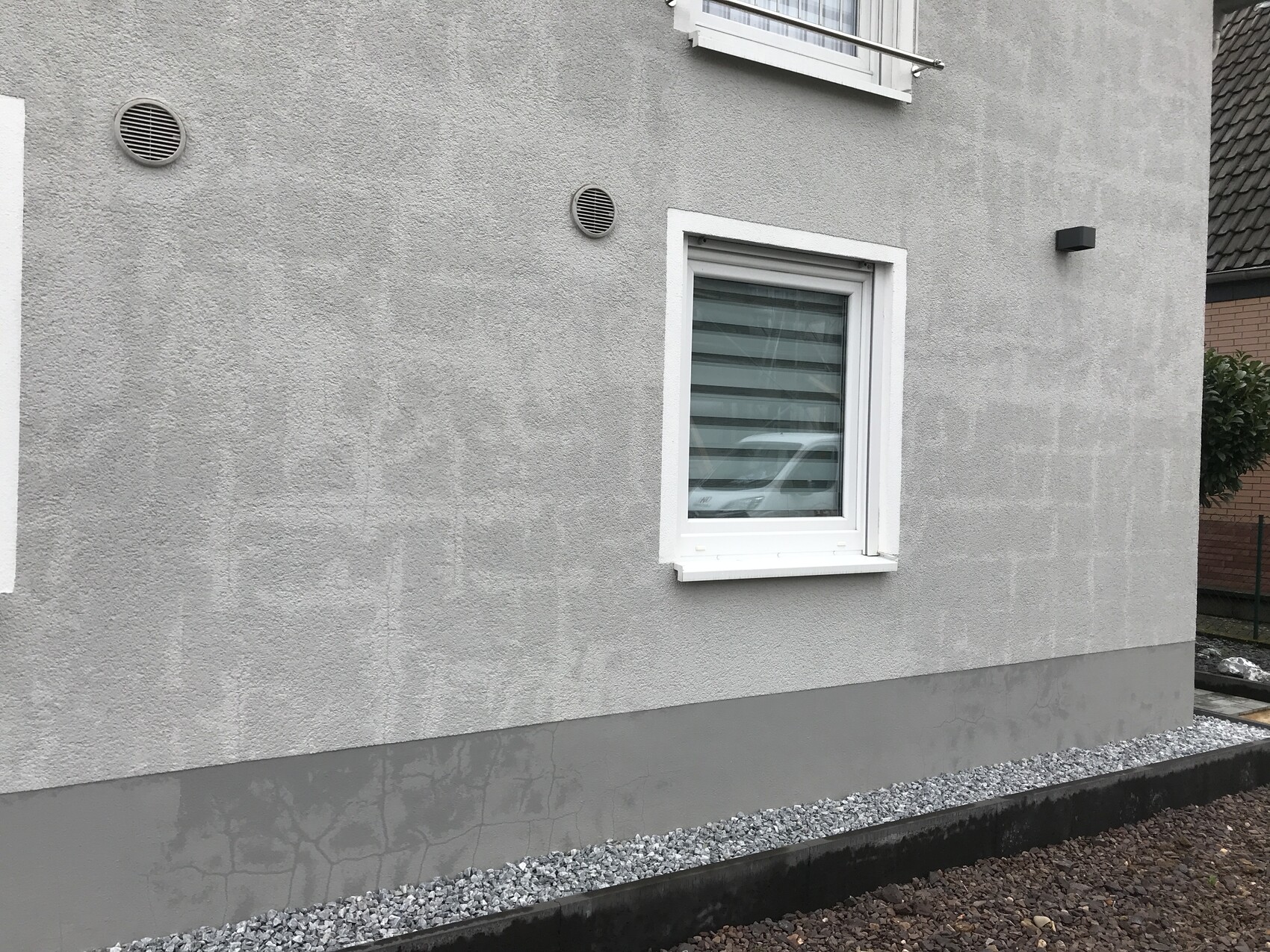 Graue Hauswand mit weißem Fensterrahmen, horizontalen Lamellen und Kiesstreifen am Fundament.