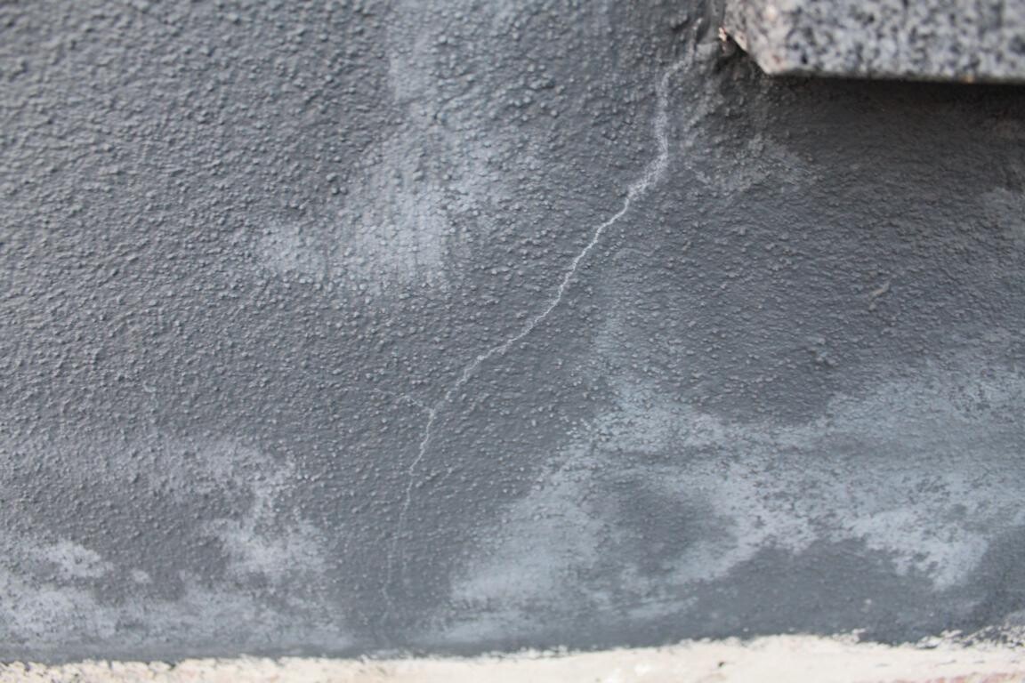 Graue, rauhe Wand mit Risslinie und Verfärbungen, unten sandfarbener Boden.