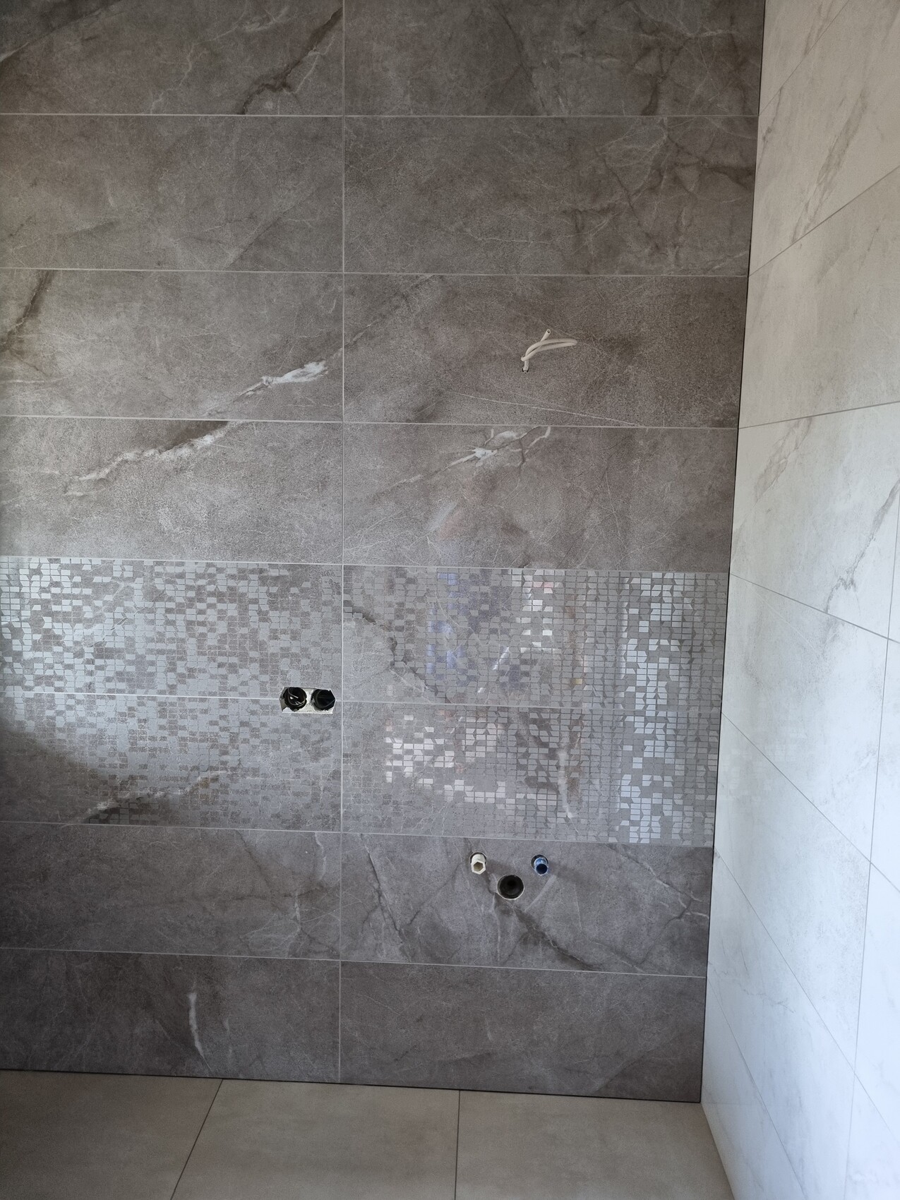 Dunkelgraue Wandfliesen mit silbernem Mosaikband; offene Rohranschlüsse der Dusche sichtbar.