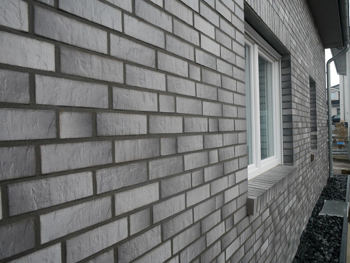 Außenansicht eines Hauses mit grauer Ziegelwand, weißen Fensterrahmen und Kies vor der Wand.