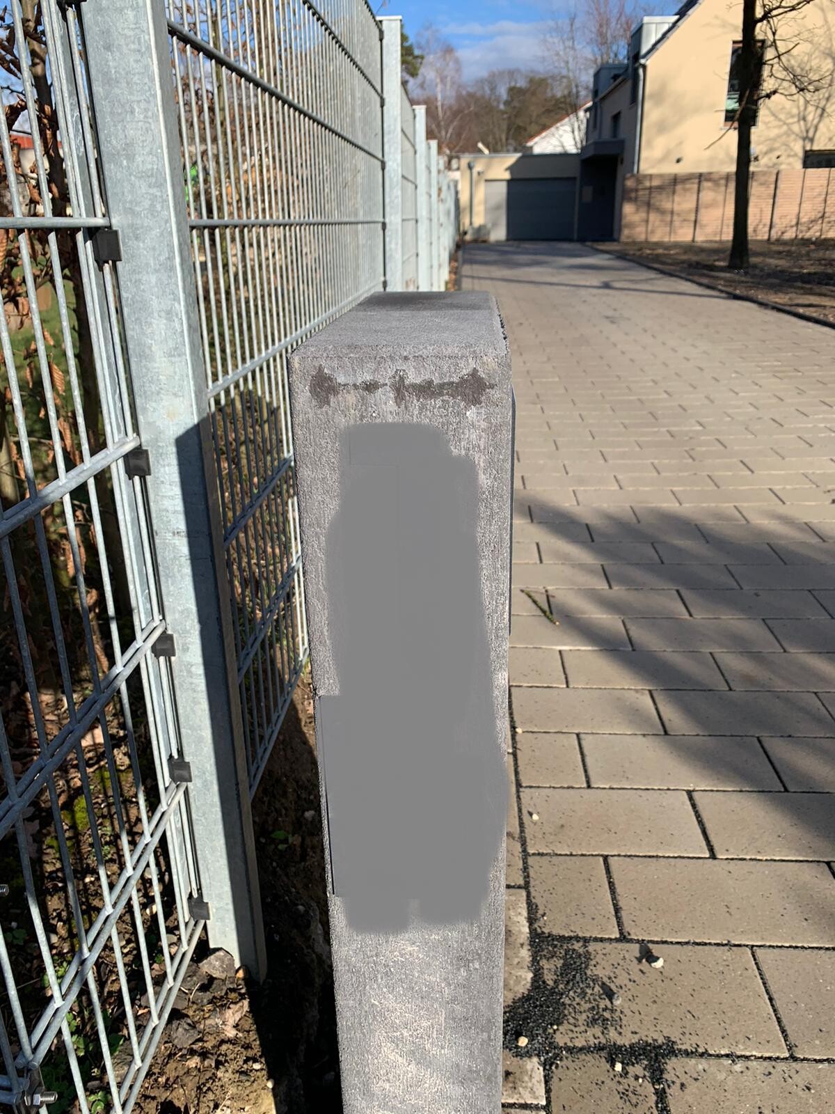Gehweg mit grauem Betonpfosten neben Metallzaun; großer grauer Fleck auf dem Pfosten.