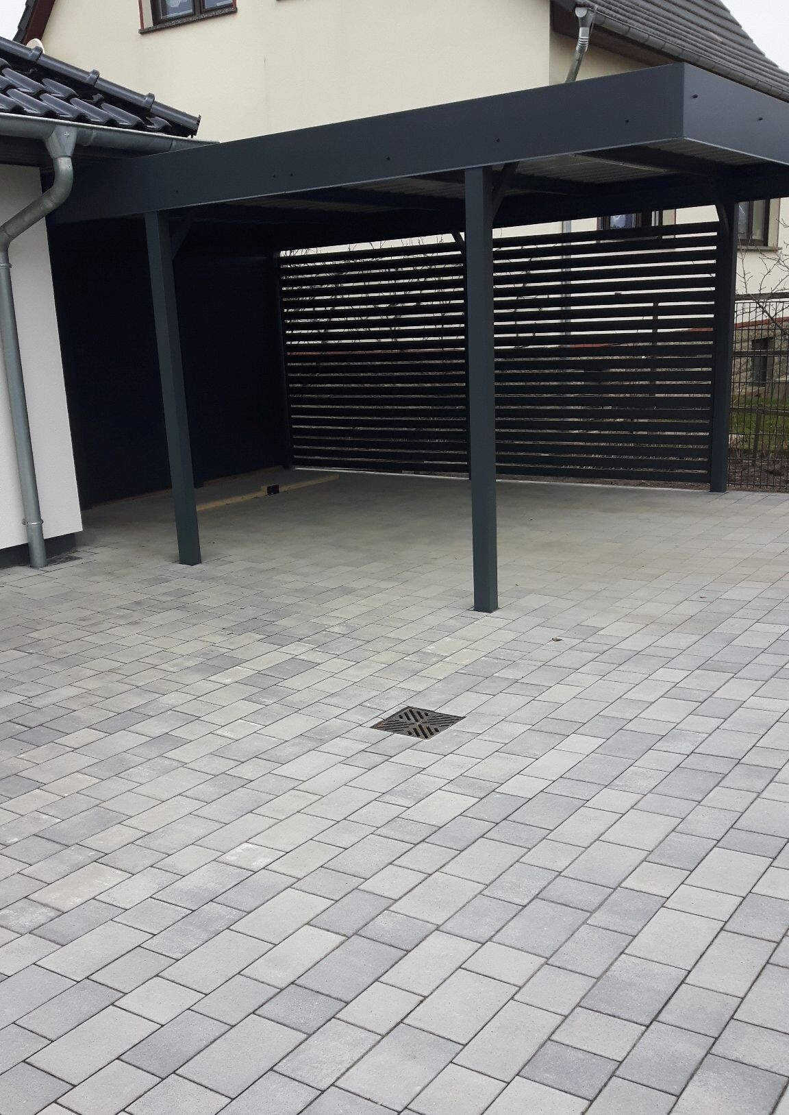 Dunkler Stahl-Carport mit horizontalen Lamellenwänden über gepflasterter Hoffläche