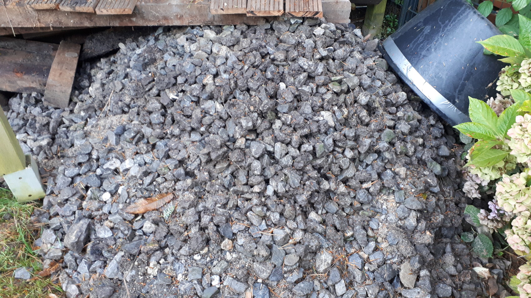 Grober grauer Schotter liegt auf dem Boden; links Holzplatten, rechts Blütensträucher am Rand.