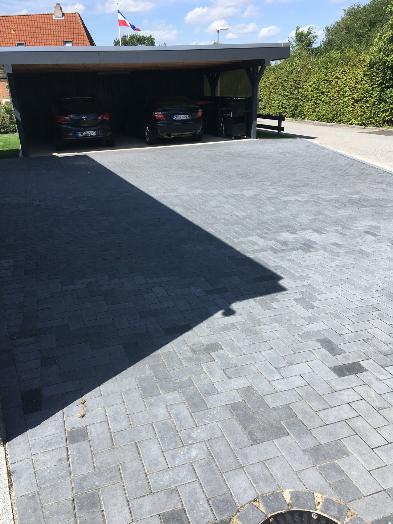Überdachter Carport mit zwei Autos auf gepflastertem Hof, grüne Hecke im Hintergrund