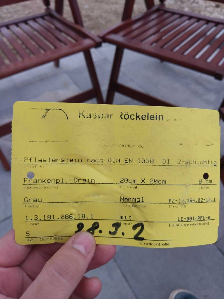Gelbe Karte mit Textdaten wird von einer Hand gehalten; im Hintergrund rote Holzstühle im Freien.