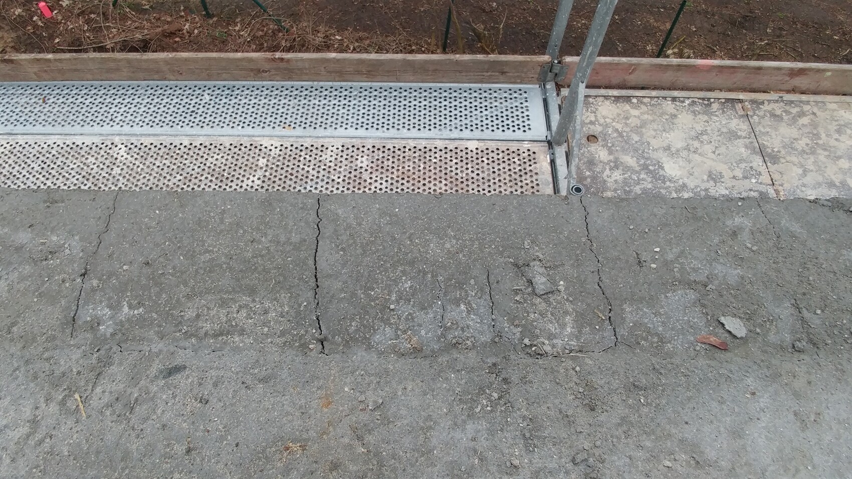 Betonboden mit Rissen; daneben perforiertes Metallraster und Geländerpfosten einer Baukonstruktion.