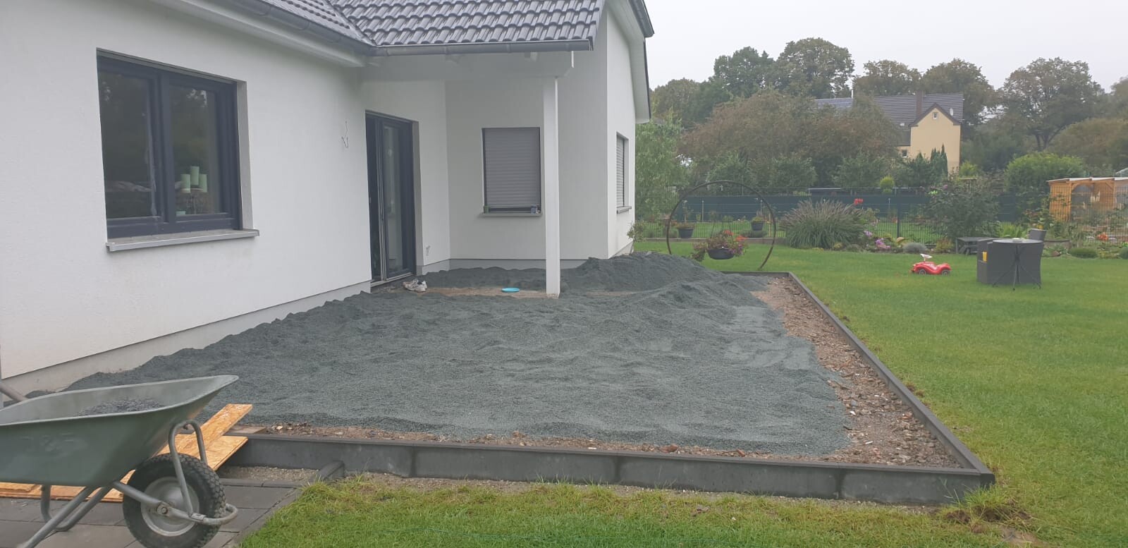 Modernes Einfamilienhaus; im Garten wird eine Kiesfläche neben dem Haus angelegt, Schubkarre links.
