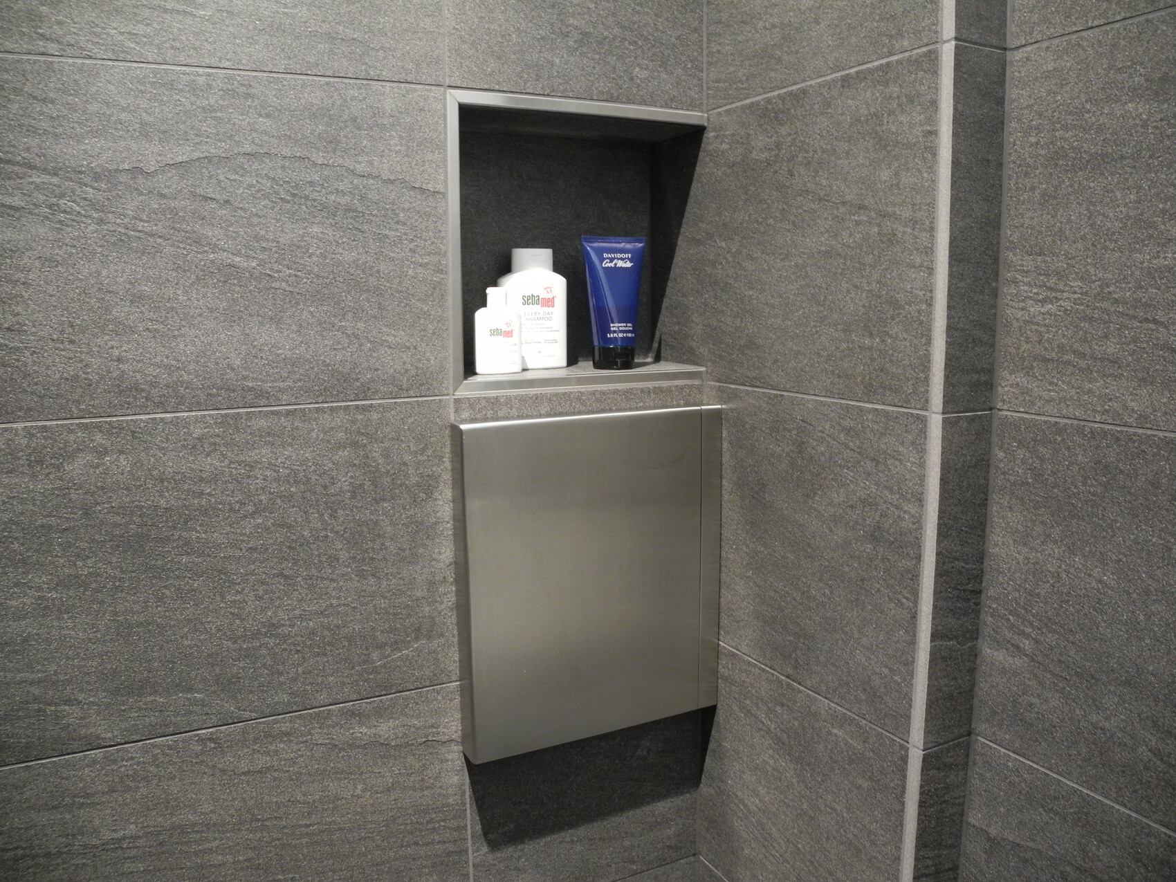 Nische in grauer Fliesenwand mit drei weißen Flaschen, blauer Tube und Edelstahlabdeckung.