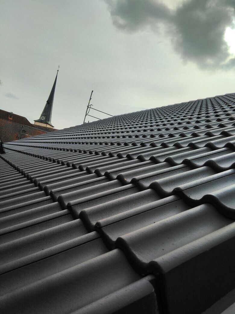Nahaufnahme eines grauen Ziegeldaches mit Kirchturm im Hintergrund und Wolkenhimmel